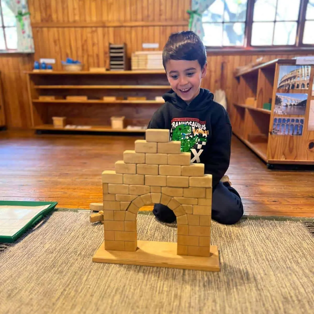 A child kneeling on a classroom mat beside the Roman arch he has built from the Montessori wooden arch material